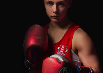 England's boxing future star, Jade Ashore wearing the red England Boxing top, emerging out of the darkness holding up her red boxing gloves. The picture looks soft and ethereal and belays the true fighting power of Jade who hopes to be at the Tokyo Olympics in 2020. Image used by Sheffield food photgrapher Mark from Wildsmith.photography's
