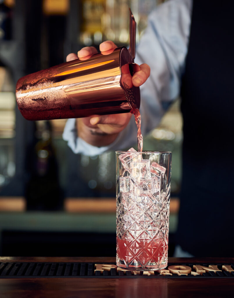 A close-up shot of a barman pouring a cocktail with a shiny brass cocktail shaker into a glass which is full of ice.Wildsmith photography commercial food photographer & food stylist based in London & Sheffield for restaurants, editorial or advertising assignment commissions. Image used by Sheffield food photgrapher Mark from Wildsmith.photography's